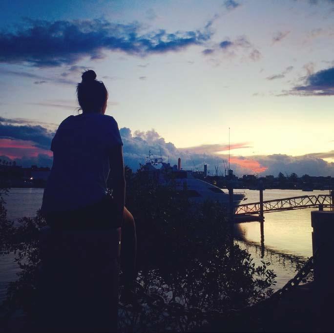 A woman is sitting on the edge of a building overlooking an inner city river as the sunsets
